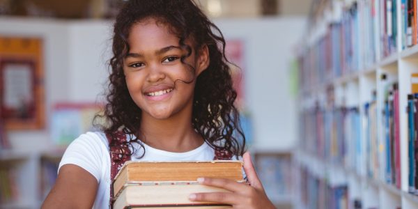portrait-of-smiling-african-american-schoolgirl-carrying-stack-of-books-in-school-library.jpg